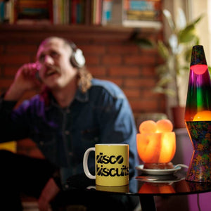 A man wearing headphones with a yellow Disco Biscuits coffee mug nearby. 