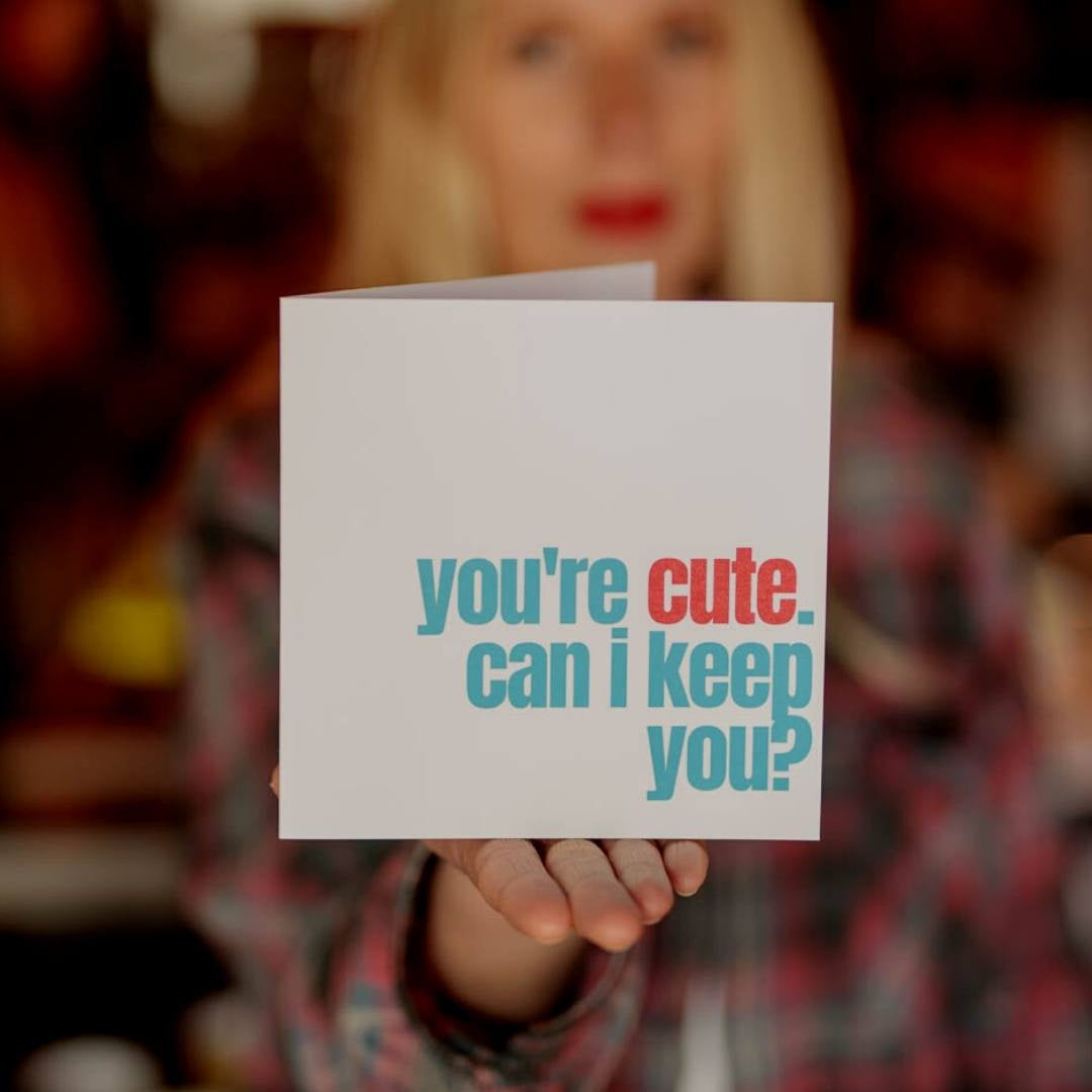 A woman holding up a notecard that says "You're cute. Can I keep you?"
